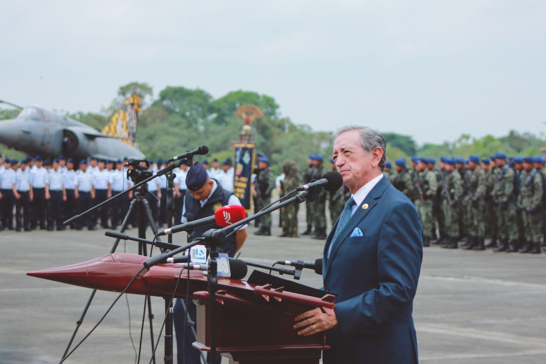 A military officer speaking at microphones during a press conference, with uniformed personnel and an aircraft in the background