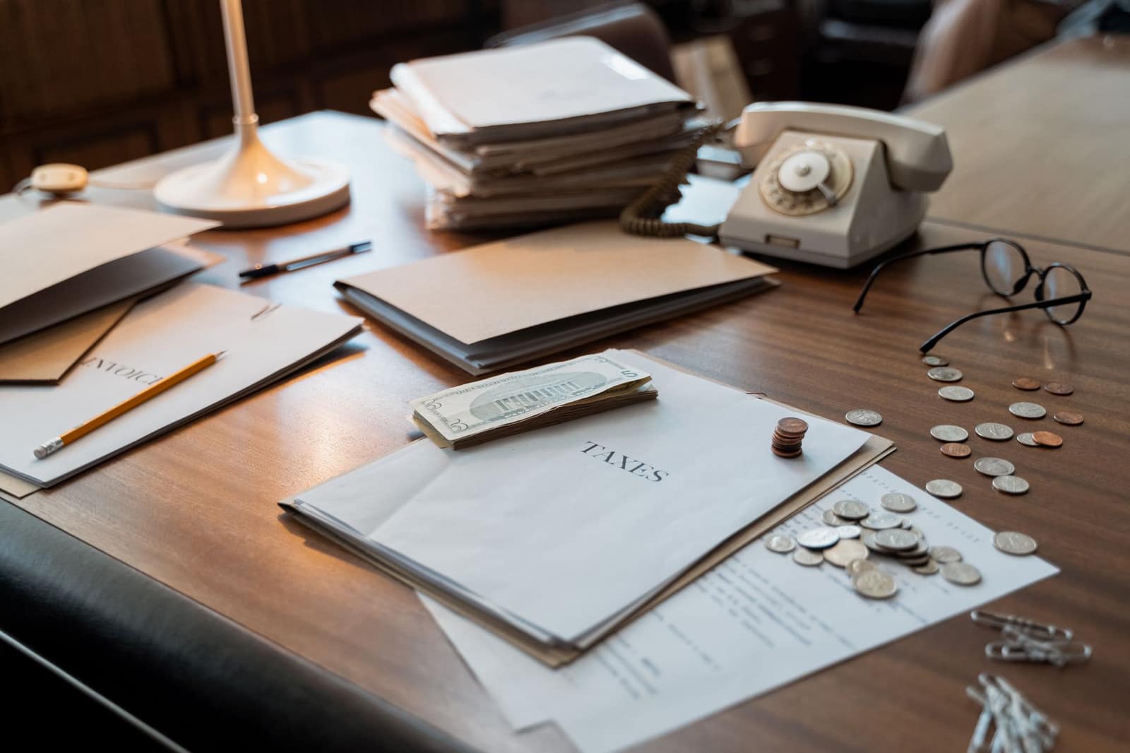 Close-up of an office desk with tax documents, coins, glasses, and an old phone