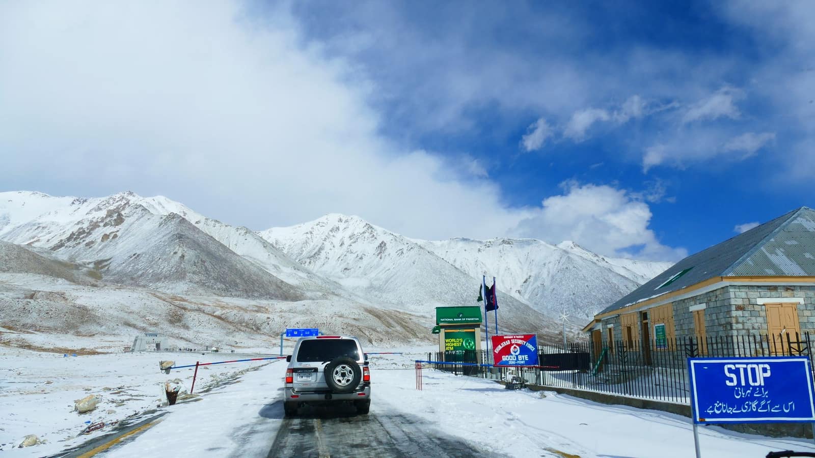 A vehicle approaches a snow-covered mountain pass with border signs and snowy peaks in the background