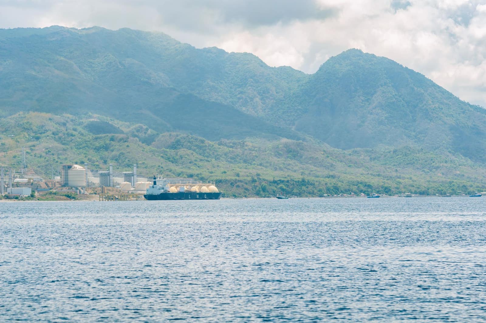 An industrial port facility with an LNG tanker docked, mountains in the background