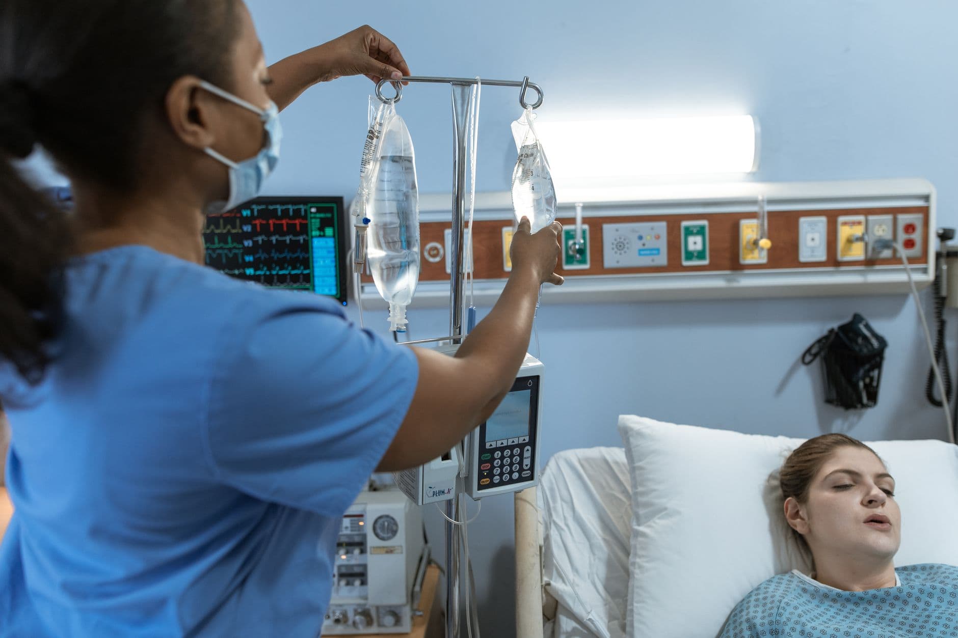 A nurse in a hospital setting adjusts an IV drip for a patient lying in bed