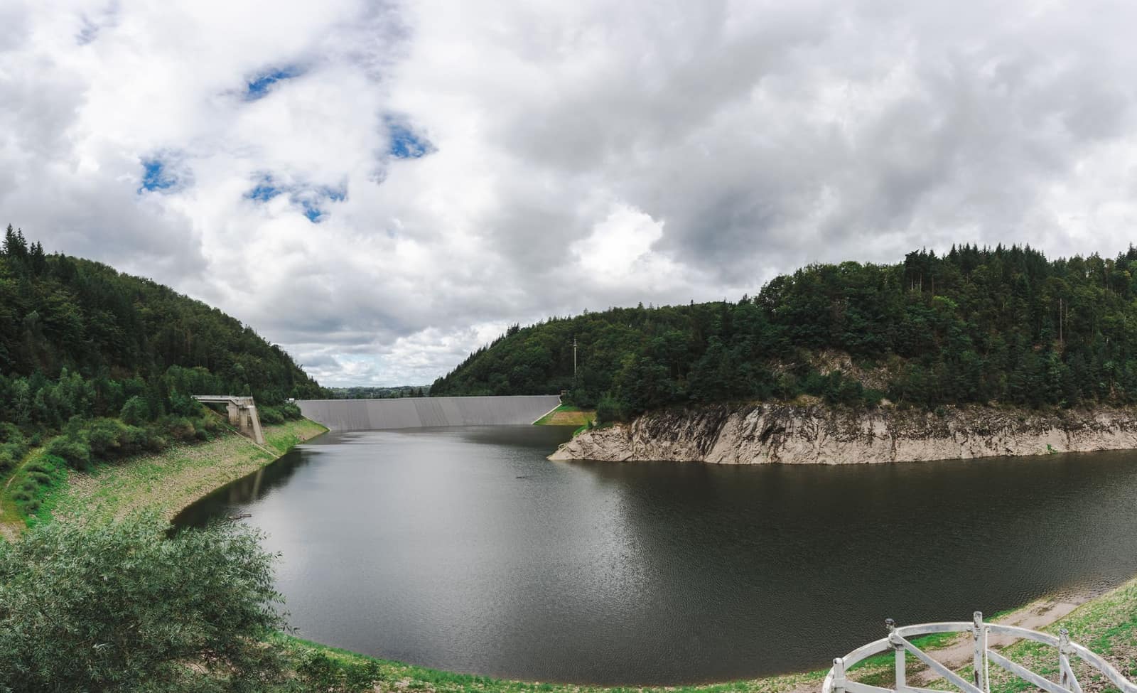 Panoramic view of a dam with a peaceful reservoir surrounded by lush green forests under a cloudy sky