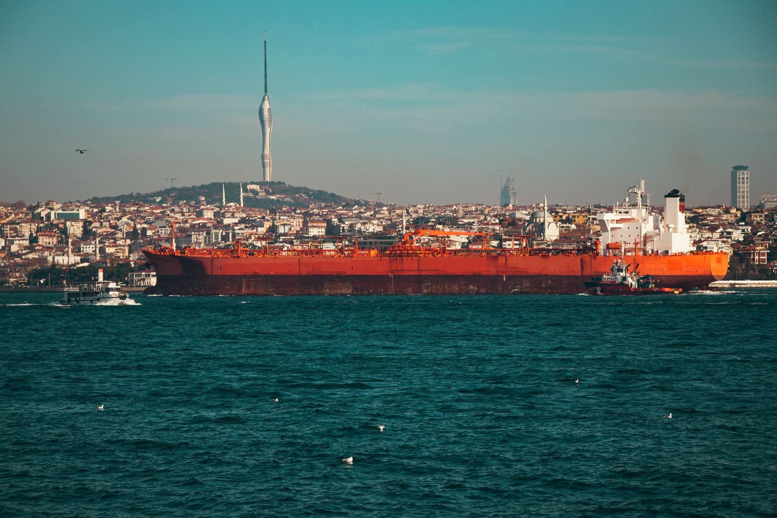 A large oil tanker navigates a strait waterway near a city skyline