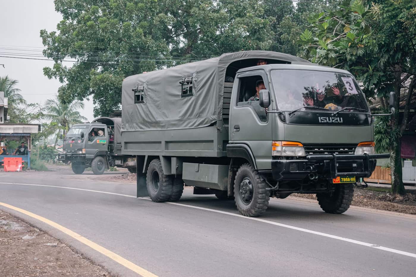 Military trucks traveling on a rural road surrounded by trees and local buildings